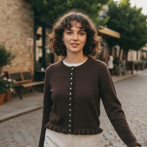Peut inclure: Un cardigan marron foncé avec des boutons nacrés et un ourlet à volants. Le pull à manches longues est porté sur un t-shirt blanc. La personne marche sur une rue pavée. Le cardigan a un col rond et une rangée de boutons.