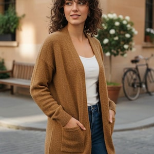 May include: A woman wearing a long, open-front, brown cardigan with two front pockets. She is also wearing a white top and blue jeans. The cardigan is a neutral color and appears to be made of a soft, textured material.