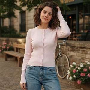 Peut inclure: Un cardigan rose clair à manches longues et à boutons. Il est porté avec un jean bleu clair. Un vélo et un banc sont en arrière-plan. La scène se déroule en extérieur.