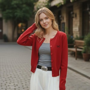 Peut inclure: Une femme porte un cardigan rouge avec un détail de nœud, un débardeur gris et un pantalon blanc. Elle porte également une ceinture noire avec une boucle argentée. Un collier complète la tenue.