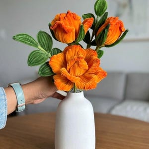 May include: A bouquet of three vibrant orange velvet flowers with ruffled petals and textured green leaves, arranged in a smooth, matte white ceramic vase with a narrow neck and a rounded base.
