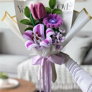 May include: A bouquet of purple and lavender pipe cleaner flowers including a tulip, a daisy, and a lily, wrapped in white paper with gold trim and the text "YOU LOVE with you". A light purple ribbon is tied around the stems.