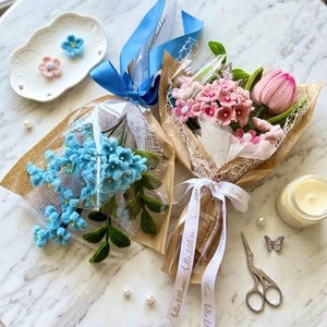 May include: Two bouquets of crocheted flowers wrapped in brown paper and white mesh. One bouquet features blue flowers with a blue ribbon, while the other contains pink flowers with a white ribbon printed with the text hello little one.