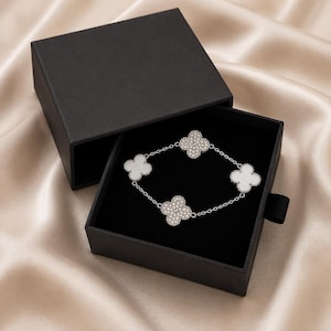 Op de afbeelding: Een zilveren armband met vier klavervormige bedels, twee met heldere kristallen en twee met wit email, ligt in een open zwarte geschenkdoos. De binnenkant van de doos is gevoerd met zwart fluweel.