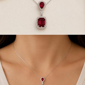May include: A silver necklace featuring a pendant with a large, rectangular, deep red gemstone surrounded by small, clear stones. Above the rectangular stone is a smaller, pear-shaped red gemstone, also encircled by clear stones. The necklace is displayed on a white background.