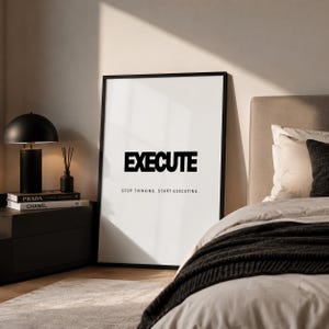 May include: A framed black and white print with the word "EXECUTE" in bold letters. Below the word is the phrase "STOP THINKING. START EXECUTING." The print is displayed in a bedroom setting, next to a black nightstand with books and a lamp.