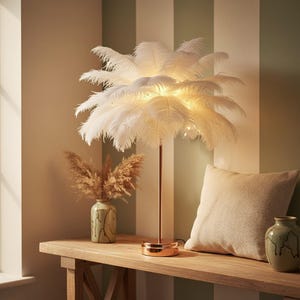 May include: A decorative table lamp with a copper-colored stem and base, topped with a cluster of white feathers. The lamp is illuminated, casting a warm glow. A vase with dried flowers and a beige pillow are also on the wooden table.