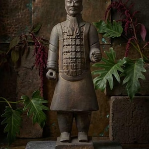 Pode incluir: Uma estátua de guerreiro em terracota cinzento-escura com uma placa peitoral detalhada, uma túnica de gola alta e um penteado com um nó. A figura assenta numa base retangular com caracteres chineses ténues gravados na extremidade frontal.