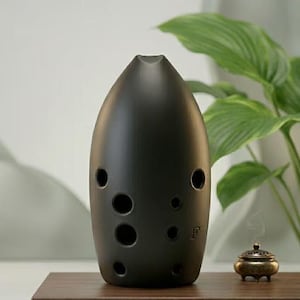 Peut inclure: Une ocarina en céramique noire mate avec un corps lisse en forme de goutte allongée, dotée de plusieurs trous pour les doigts et de la lettre F en relief sur le côté inférieur droit. L'instrument possède un embout et une base arrondie.