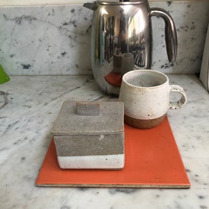 May include: A silver French press coffee maker, a white ceramic mug, and a white ceramic box with a lid, all sitting on a bright orange square tile.