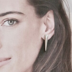 Könnte beinhalten: Ein silberfarbener, geometrischer Ohrstecker mit dreieckiger Form. Der Ohrring wird am Ohr getragen, mit einem nach unten gerichteten, spitzen Design. Der Ohrring ist ein schlichtes, modernes Accessoire.