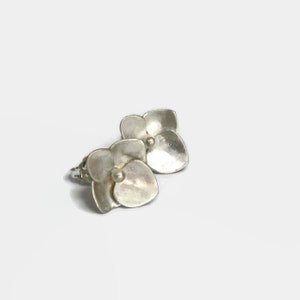 Peut inclure: Une paire de boucles d'oreilles argentées en forme de fleur. Chaque boucle d'oreille présente un groupe de pétales avec un petit accent rond au centre. Les boucles d'oreilles sont posées sur un fond blanc.