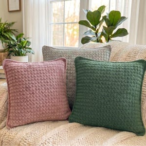 May include: Three square throw pillows in shades of pink, gray, and green rest on a cream-colored knitted blanket. The pillows have a textured, woven appearance and are arranged on a sofa near a window and potted plants.