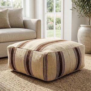 May include: A square floor pouf featuring a textured beige fabric base with prominent vertical dark brown stripes. The ottoman has a low-profile, boxy shape with defined edges and a woven appearance, providing a firm surface for seating or foot support.
