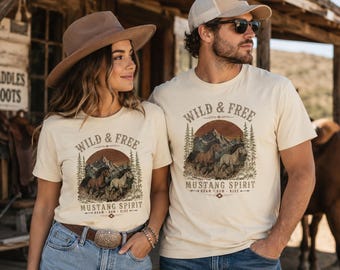 Puede incluir: Dos personas vistiendo camisetas color crema con un diseño gráfico. El diseño presenta el texto "WILD & FREE" sobre una escena de montaña con caballos y las palabras "MUSTANG SPIRIT". Las camisetas se combinan con jeans y un cinturón marrón.