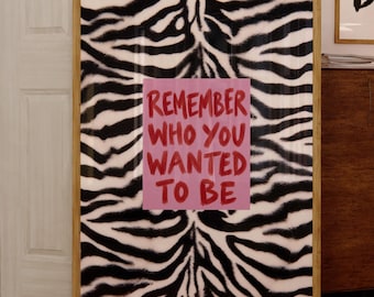 Peut inclure: Une œuvre d'art murale rectangulaire encadrée présentant un motif zébré noir et blanc. Au centre, un carré rose contient le texte rouge : REMEMBER WHO YOU WANTED TO BE. Le cadre est en bois clair et est posé à la verticale.
