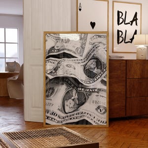 Op de afbeelding: Een rechthoekig ingelijst kunstwerk aan de muur met een zwart-wit fotoprint van meerdere verfrommelde Amerikaanse dollarbiljetten. Het kunstwerk is ingelijst in een lichtgekleurde houten lijst, die de gedetailleerde textuur van het papiergeld laat zien.