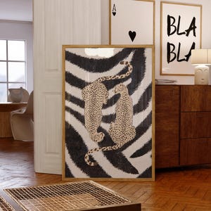 Op de afbeelding: Een rechthoekige ingelijste kunstprint voor aan de muur met twee gevlekte cheeta's tegen een achtergrond met opvallende zwart-witte zebrastrepen. Het kunstwerk zit in een licht houten frame en toont een contrastrijk dierenmotief.