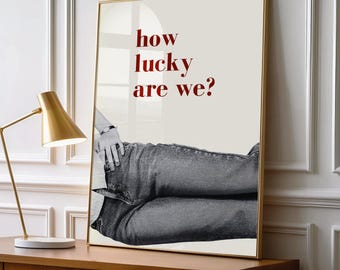 Peut inclure: Une affiche murale rectangulaire dans un cadre doré présentant une photographie en noir et blanc d'une personne portant un jean, avec le texte rouge "how lucky are we?" imprimé sur un fond beige clair au-dessus de l'image du jean.