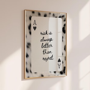 May include: A rectangular wall art piece featuring an Ace of Hearts playing card design with a black and white animal print border. The center text reads, "risk is always better than regret" in black cursive script, enclosed in a thin gold frame.