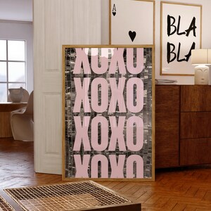 May include: A rectangular framed wall art piece featuring the repeating text XOXO in large, light pink capital letters. The background behind the letters consists of a vertical, textured pattern of alternating black and white brushstrokes.