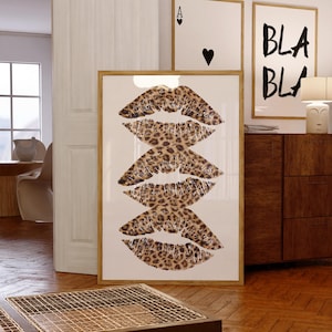 May include: A rectangular framed wall art print featuring a vertical stack of three lip-shaped graphics with a brown and black leopard print pattern on a white background. The frame is made of light-colored wood and is positioned on a wooden floor.
