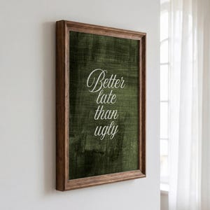 May include: A rectangular wall art piece in a dark brown wooden frame. The print features a dark olive green textured background with the white cursive text Better late than ugly centered on the surface. The frame has a simple, clean profile.