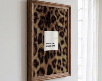 Puede incluir: Una pieza de arte mural rectangular en un marco de madera marrón oscuro con un fondo de estampado de leopardo. Centrado en el estampado hay un cuadrado blanco que contiene el texto negro "WHAT IF IT ALL WORKS OUT?" en una fuente sans-serif.