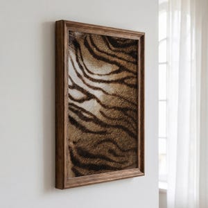 May include: A rectangular wall art piece featuring a close-up print of tiger fur with distinct black stripes on a golden-brown background, encased in a dark brown wooden frame with a beveled edge, displayed at an angle on a plain white wall.
