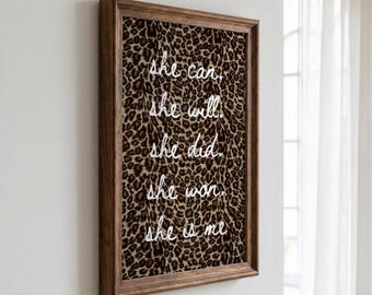Peut inclure: Une œuvre d'art murale rectangulaire dans un cadre en bois brun foncé avec un fond à motif léopard. Le texte blanc en cursive indique : she can. she will. she did. she won. she is me. L'œuvre est présentée en angle sur un mur uni.