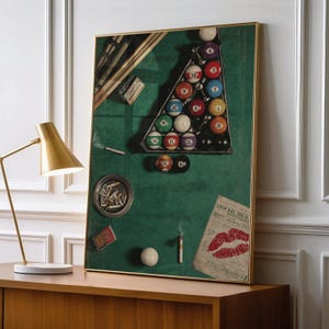 May include: A framed wall art print depicting a green pool table surface with a triangular rack of numbered billiard balls, pool cues, a full ashtray, a pack of cigarettes, a single lit cigarette, and a paper note featuring a red lip print.