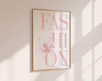 May include: A rectangular wall art print in a thin gold frame featuring the word FASHION in large, light pink capital letters. A single, soft pink lily flower is positioned over the letters, partially obscuring the middle section of the text.