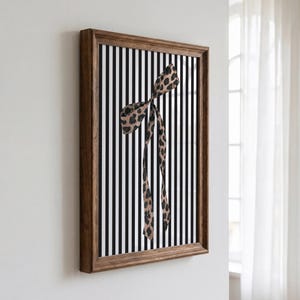 May include: A rectangular wall art piece in a dark brown wooden frame featuring a vertical black and white striped background with a central leopard print ribbon tied in a bow with two long, dangling ends hanging down the centre of the frame.