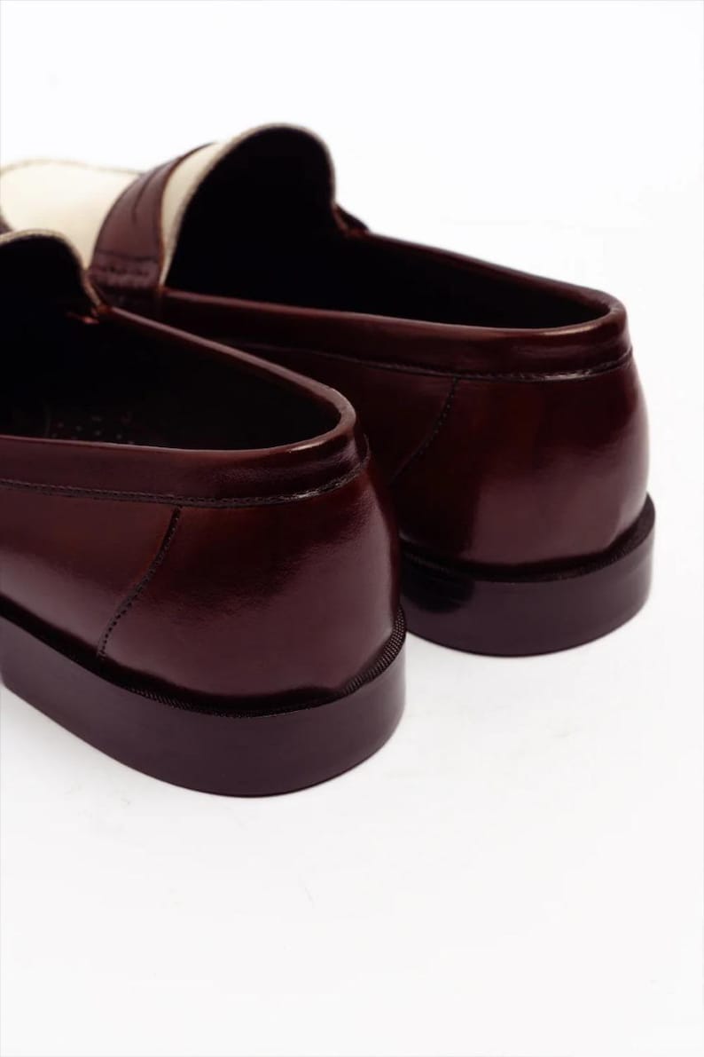 Pu&ograve; includere: Primo piano di due mocassini in pelle marrone con un accento bianco sulla parte superiore. Le scarpe hanno una finitura lucida e una suola marrone scuro. L'immagine &egrave; su sfondo bianco, evidenziando le calzature.