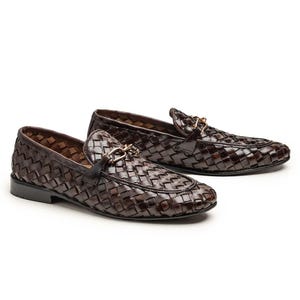 May include: A pair of dark brown loafers featuring a woven leather upper and a gold-toned metal horsebit detail across the vamp. The shoes have a low heel and a rounded toe, showcasing a consistent basket-weave pattern throughout the exterior.