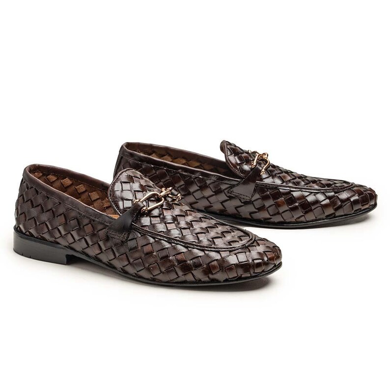 May include: A pair of dark brown loafers featuring a woven leather upper and a gold-toned metal horsebit detail across the vamp. The shoes have a low heel and a rounded toe, showcasing a consistent basket-weave pattern throughout the exterior.