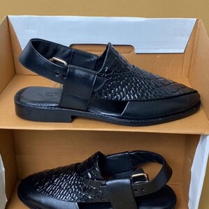 Pode incluir: Sand&aacute;lias de couro preto com textura tecida na parte superior e fecho de fivela. As sand&aacute;lias s&atilde;o exibidas em uma caixa de papel&atilde;o, mostrando seu design e estilo. O cal&ccedil;ado &eacute; adequado para uso casual.