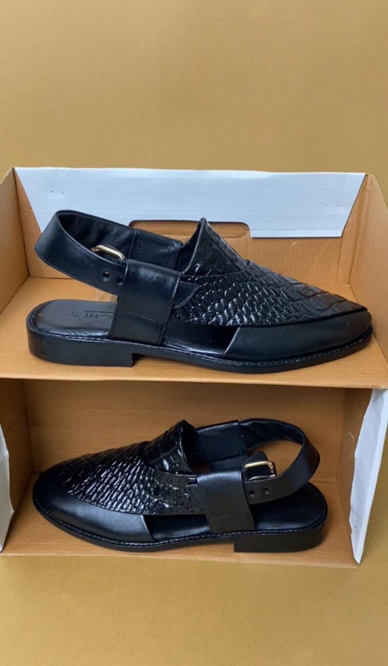 Pode incluir: Sand&aacute;lias de couro preto com textura tecida na parte superior e fecho de fivela. As sand&aacute;lias s&atilde;o exibidas em uma caixa de papel&atilde;o, mostrando seu design e estilo. O cal&ccedil;ado &eacute; adequado para uso casual.