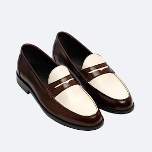 Pu&ograve; includere: Un paio di mocassini penny in pelle marrone e bianca. Le scarpe hanno un design classico con tomaia marrone e mascherina bianca. Le suole sono nere.