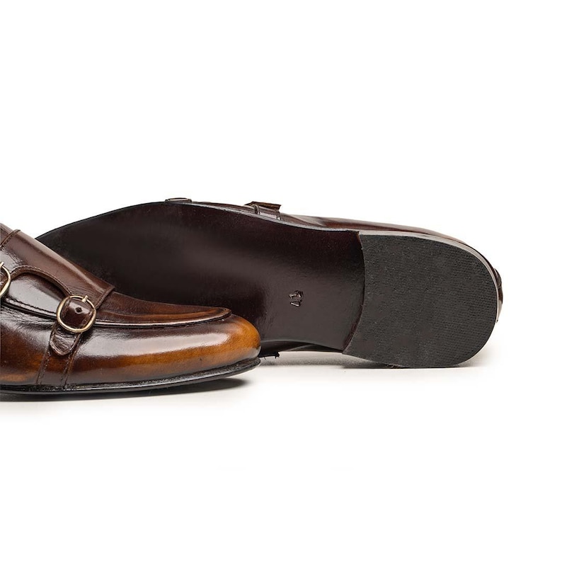 Op de afbeelding: Een paar donkerbruine leren schoenen met dubbele gesp en een gepolijste afwerking. De schoenen zijn voorzien van twee goudkleurige metalen gespen aan de zijkant en een gladde, donkerbruine zool. Het leer heeft een subtiel kleurverloop.