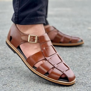 May include: A pair of brown leather fisherman sandals featuring a woven strap design, a closed toe, and an adjustable ankle strap with a gold-tone metal buckle. The sandals have a dark sole with a thin tan layer along the edge of the base.