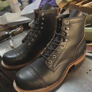 Puede incluir: Un par de botas de cuero negro con puntera reforzada y perforaciones decorativas tipo brogue, ojales de metal plateado y cordones oscuros. Las botas tienen una entresuela de cuero marrón claro visible con construcción de vira cosida.