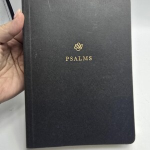 May include: A black hardcover book with a textured finish. The front cover features the gold-colored ESV logo at the top center, followed by the word PSALMS printed in gold capital letters directly below the logo in a simple serif font.