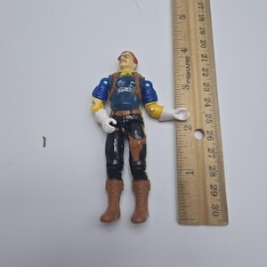 May include: A vintage action figure of a man in a blue shirt, black pants, and brown boots. The figure has a red head, yellow scarf, and white gloves. A wooden ruler is next to the figure.