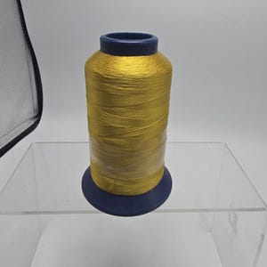 May include: A spool of vibrant golden yellow embroidery thread wound tightly around a cylindrical core. The spool features a dark blue plastic top and base, with a clear protective plastic film wrapped around the center of the thread.