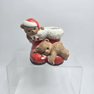 May include: A ceramic Christmas decoration featuring a red boot with white trim, holding two brown teddy bears. One bear wears a Santa hat. The bears are holding candy canes. The boot is on a clear acrylic stand.