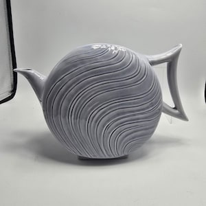 May include: A gray ceramic teapot with a spherical body and a curved handle. The teapot features a spout and is decorated with a wave pattern. The teapot is a decorative item for the kitchen or dining room.