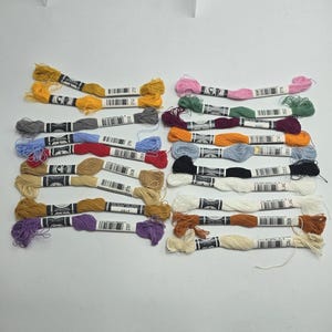 May include: A collection of twenty-two skeins of embroidery floss arranged in two columns. Each skein features a paper label with a barcode and color number. The threads display a variety of colors including yellow, gray, blue, red, brown, purple, pink, green, maroon, orange, black, white, and cream.