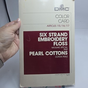 May include: A rectangular DMC color card for embroidery floss and pearl cottons. The off-white cover features the text DMC COLOR CARD ARTICLES 115/116/117, SIX STRAND EMBROIDERY FLOSS, MOULINÉ SPÉCIAL AND PEARL COTTONS, COTON PERLE, and COLOR CARD NO. W-200A 1987-1.