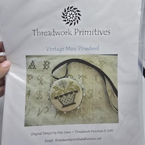 May include: A package for a cross-stitch project titled "Vintage Mini Pinwheel" by Threadwork Primitives. The design features a round, embroidered pendant with a flower pot design. The package includes the designer's email address.